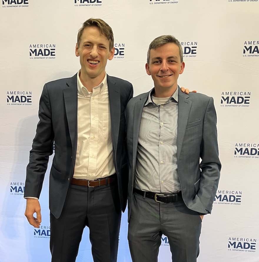 two male students standing in front of of white back drop with blue text reading "American Made"