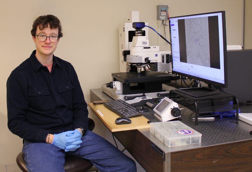 Student facing camera sitting at desk with microscope and computer