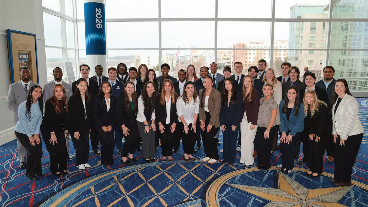 38 people standing in two rows in front of large windows at a conference center