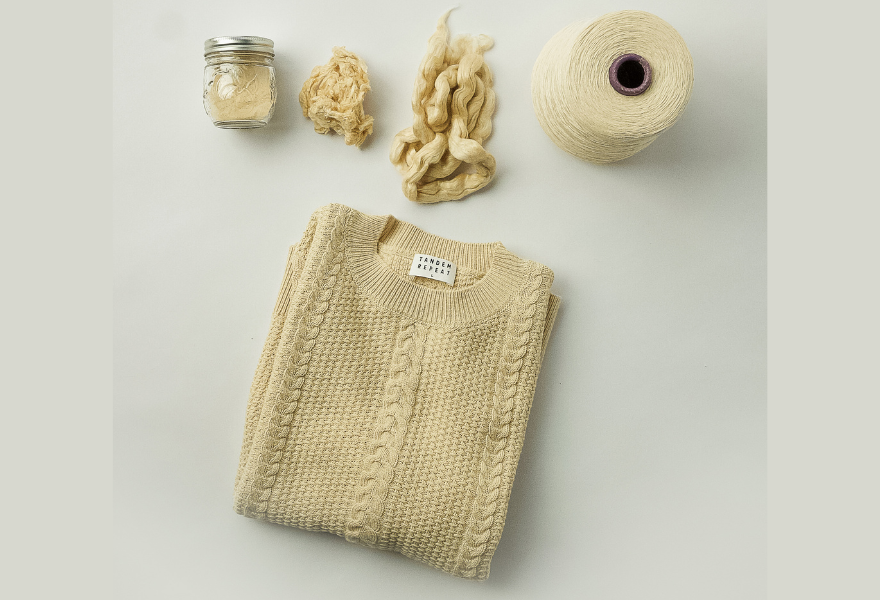 A cream-colored sweater displayed with raw fiber, a yarn cone, and a small jar, arranged in a flat lay on a light background.