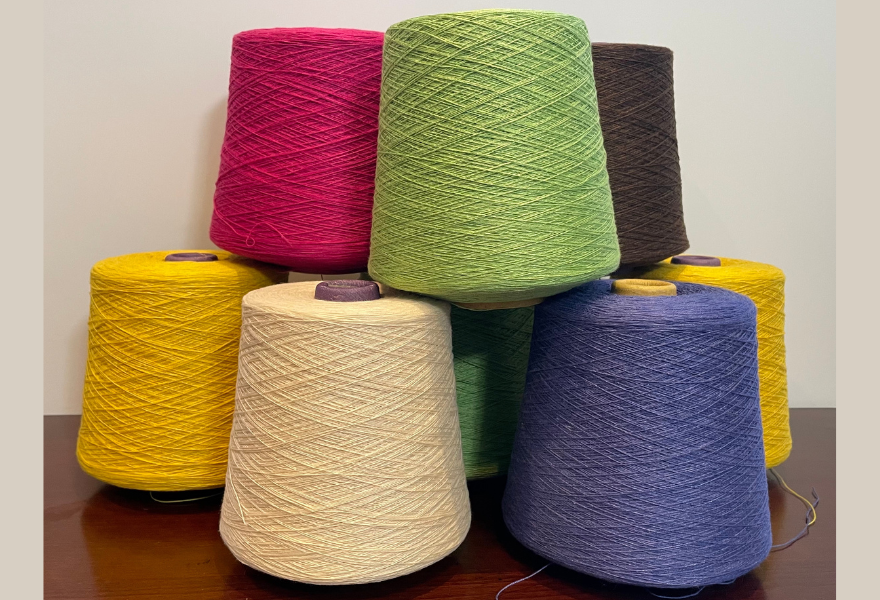 Several large cones of yarn in bright colors—magenta, light green, dark brown, yellow, cream, and blue—arranged on a table against a plain wall.