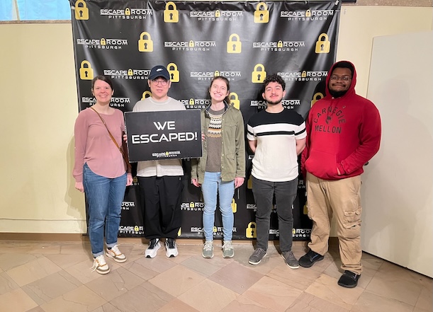Group of five people standing in front of Pittsburgh Escape Room sign with one person holding 'we escaped' sign